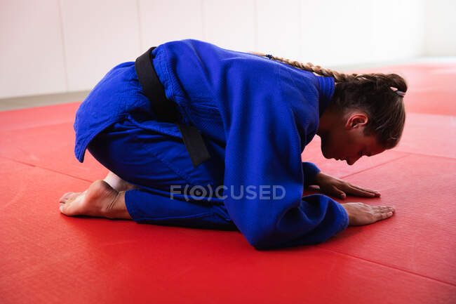 Вид сбоку на юную кавказскую дзюдоистку в синих дзюдоистах, стоящую на коленях и кланяющуюся на ковриках в тренажерном зале перед тренировкой по дзюдо. — стоковое фото