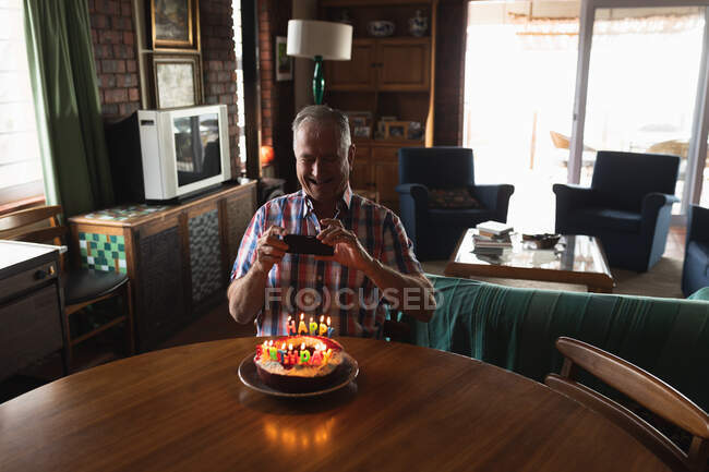Vista frontal de um homem caucasiano sênior em casa, sentado sozinho na mesa de jantar e sorrindo, tirando foto de um bolo de aniversário com velas acesas na frente dele — Fotografia de Stock