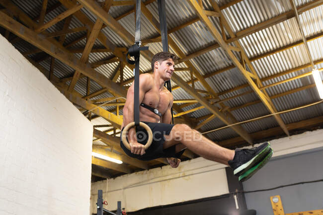 Vista lateral de un hombre atlético caucásico sin camisa con una correa en el pecho monitor de frecuencia cardíaca entrenamiento cruzado en un gimnasio, levantándose sobre anillos gimnásticos con las piernas hacia arriba - foto de stock