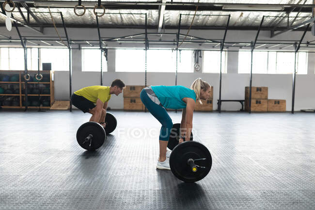Side view of an athletic Caucasian man and woman wearing sports clothes cross training at a gym, weight training with barbells, standing and bending down to lift the weights from the floor — Stock Photo