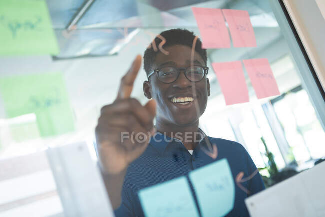 Un hombre de negocios afroamericano con una camisa azul y gafas, trabajando en una oficina moderna, escribiendo en un tablero claro con notas y sonriendo - foto de stock