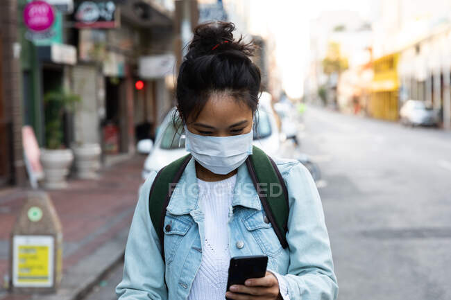 Vue de face d'une femme métisse aux longs cheveux foncés dans les rues de la ville pendant la journée, portant un masque facial contre la pollution atmosphérique et le coronavirus, debout et utilisant un smartphone avec des bâtiments en arrière-plan. — Photo de stock