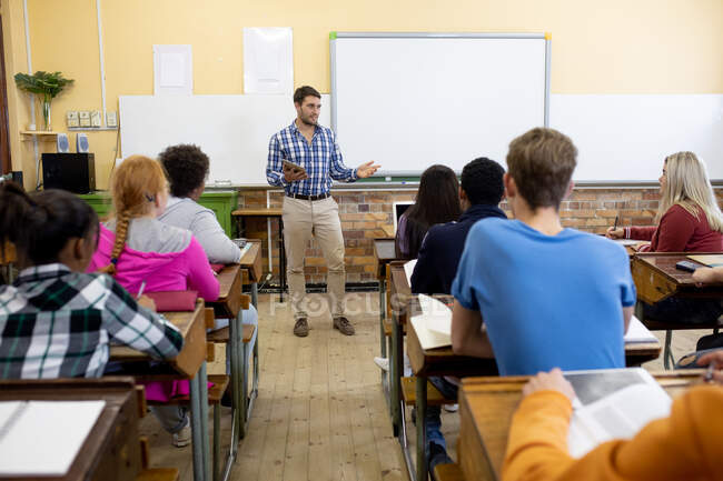 Vista frontal de un maestro caucásico de pie, sosteniendo una tableta y hablando con un grupo multiétnico de adolescentes en una escuela secundaria sentados en escritorios y escuchando - foto de stock