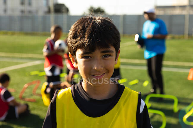 Portrait d'un jeune footballeur de race mixte, debout, regardant vers la caméra et souriant sur un terrain de jeu au soleil, avec ses coéquipiers et leur entraîneur en arrière-plan — Photo de stock