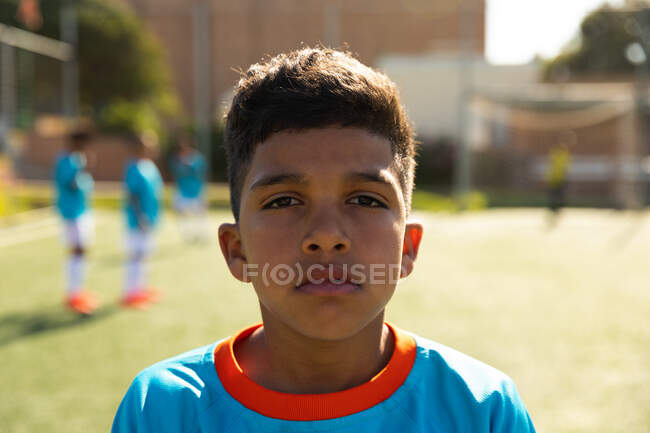 Portrait of a mixed race boy soccer player wearing a blue team strip, standing on a playing field with on a sunny day, looking to camera and smiling, with teammates in the background — Stock Photo