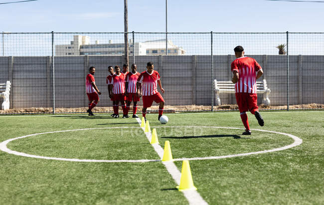 Multi ethnic team of male five a side football players wearing a team strip training at a sports field in the sun, warming up tackling with ball between cones. — Stock Photo