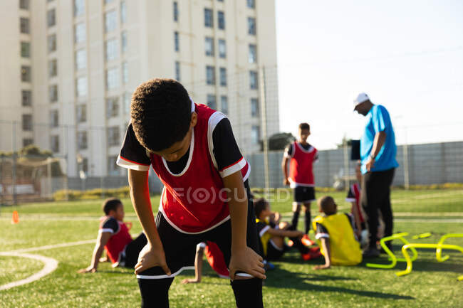 Vista frontal de un futbolista de raza mixta de pie con las manos de rodillas en un campo de juego descansando durante el entrenamiento, con sus compañeros de equipo sentados y escuchando a su entrenador en el fondo - foto de stock