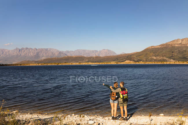 Visão traseira de um casal caucasiano se divertindo em uma viagem às montanhas, de pé em um caminho, em uma costa, uma mulher está apontando para algo, em um dia ensolarado. — Fotografia de Stock