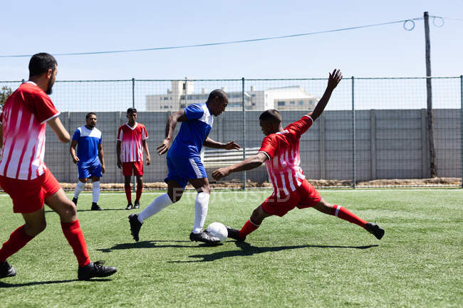 Duas equipes multi étnicas do sexo masculino cinco jogadores de futebol de um lado vestindo uma tira de equipe jogando um jogo em um campo de esportes ao sol, atacando e chutando bola . — Fotografia de Stock