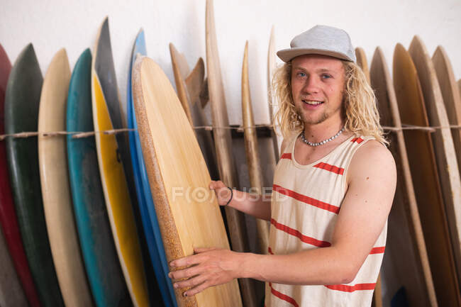 Caucásico fabricante de tablas de surf masculino en su estudio, sosteniendo una de las tablas de surf y sonriendo a la cámara, con otras tablas de surf en un estante en el fondo. - foto de stock