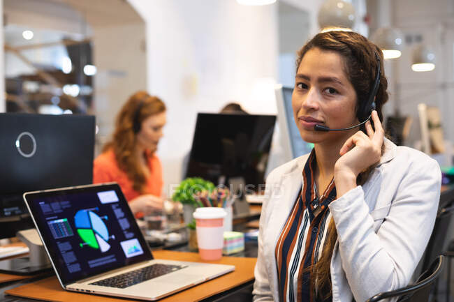 Portrait of a mixed race female business creative working in a casual modern office, talking on a phone headset and looking at camera, with colleagues working in the background — Stock Photo