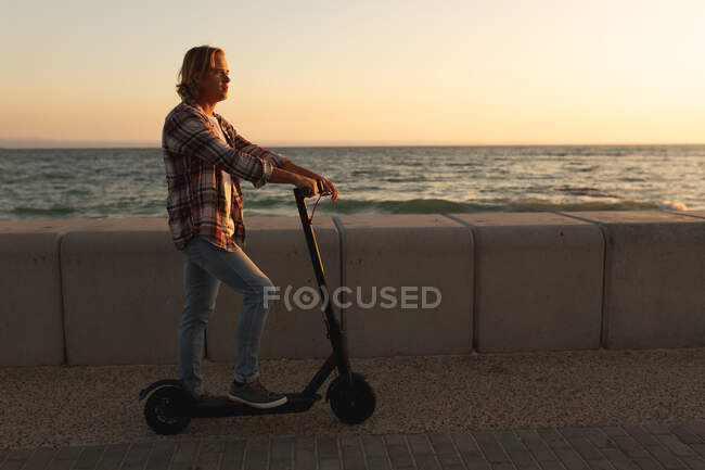 Homem caucasiano montando um e-scooter em um passeio marítimo ao pôr-do-sol, relaxando durante umas férias ativas na praia à beira-mar — Fotografia de Stock