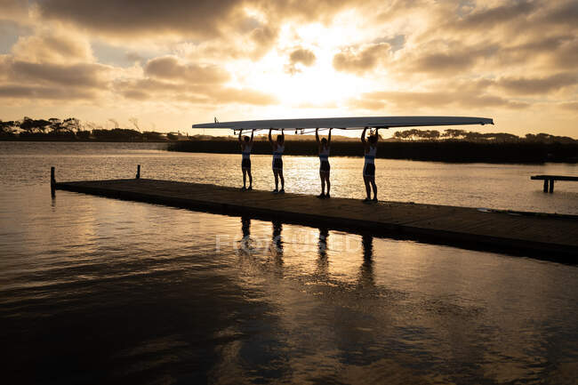 Біля підніжжя видніється веслувальний колектив з чотирьох кавказьких чоловіків, які несуть човен над головою з піднятими руками, йдучи по пристані на річці при заході сонця. — стокове фото
