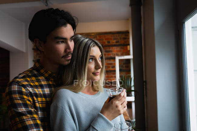 Вид сбоку на молодого человека смешанной расы и молодую кавказскую женщину, наслаждающуюся временем дома, стоящую у окна, обнимающую и пьющую кофе. — стоковое фото