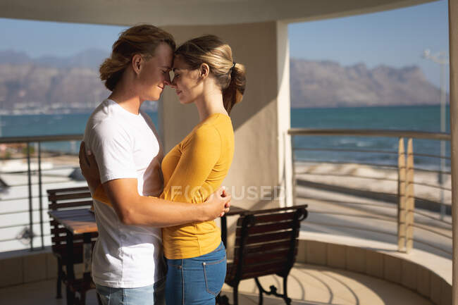 Coppia caucasica in piedi su un balcone, abbracciandosi e toccandosi la fronte. Distanziamento sociale e autoisolamento in quarantena. — Foto stock