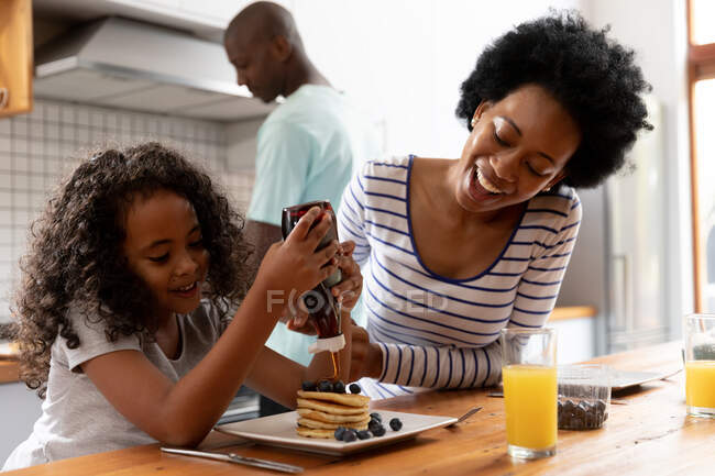 Front view close up of a young African American girl and her mother at home in the kitchen in the morning, sitting at the kitchen island, the girl pouring sauce on her pancakes, with the father standing in the background cooking — Stock Photo