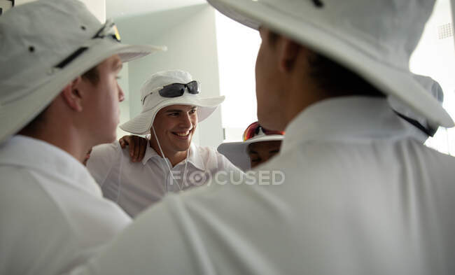 Side view close up of a group of teenage multi-ethnic male cricket players wearing whites, huddling in a changing room, embracing and smiling. — Stock Photo