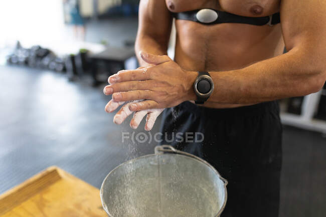 Front view mid section of a shirtless athletic man wearing a chest strap heart rate monitor cross training at a gym, rubbing his hands with chalk in preparation for weight training — Stock Photo