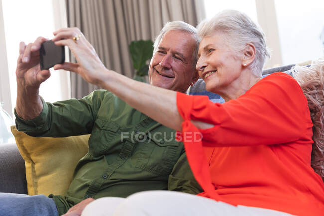 Großaufnahme eines glücklichen Rentnerehepaares aus dem Kaukasus zu Hause in ihrem Wohnzimmer, auf einem Sofa sitzend, das Smartphone zusammenhaltend, beide auf das Telefon blickend, ein Selfie machend und lächelnd, Paar isolierend während der Coronavirus-Pandemie 19 — Stockfoto