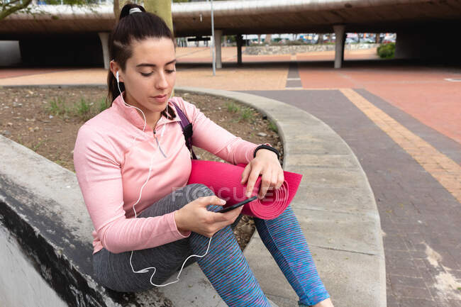 Вид спереди на белую женщину, идущую на тренировку, с спортивной сумкой и ковриком для йоги, с помощью смартфона и наушников, сидящую на стене в парке — стоковое фото