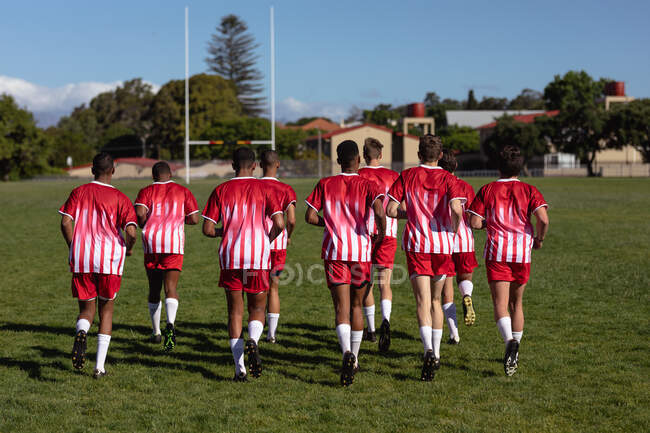 Vista trasera de un equipo masculino multiétnico adolescente de jugadores de rugby que usan su tira de equipo, corriendo juntos en el campo de juego. - foto de stock