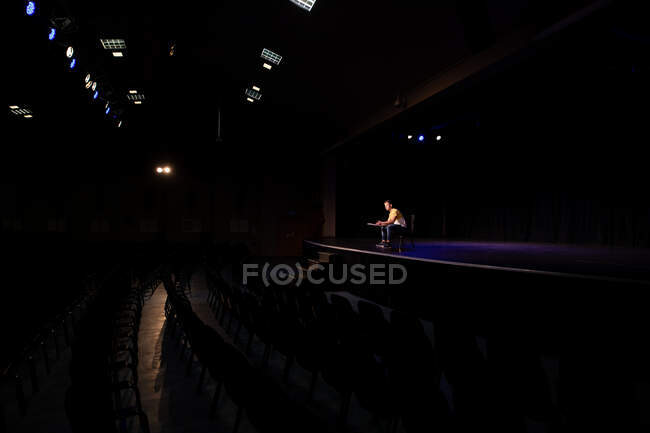 Vista lateral lejana de un adolescente caucásico estudiante de secundaria en un teatro de secundaria vacío preparándose antes de una actuación, sentado en la silla en el escenario, sosteniendo un guion y practicando su parte - foto de stock