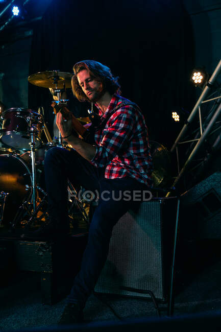 Vista lateral de cerca de un guitarrista caucásico ensayando en un lugar de música antes de una actuación, sentado en un amplificador, concentrando y tocando su guitarra eléctrica - foto de stock