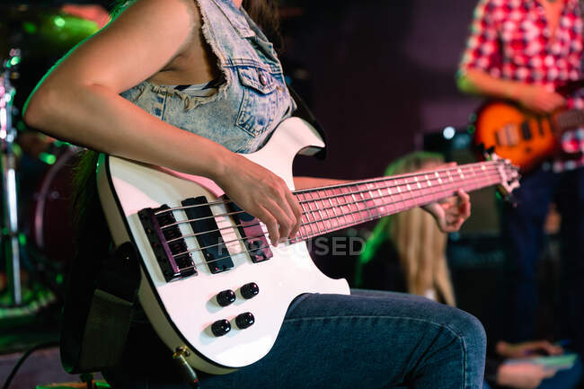 Вид сбоку на среднюю часть бас-гитаристки, репетирующей в музыкальном зале перед выступлением, сидящей и играющей на белой бас-гитаре с другим музыкантом на заднем плане — стоковое фото