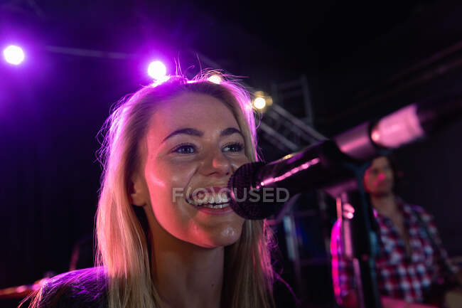 Vue de face gros plan d'une chanteuse caucasienne jouant dans un lieu de musique avec un groupe, souriant et chantant dans un microphone, avec des lumières roses et un musicien en arrière-plan — Photo de stock