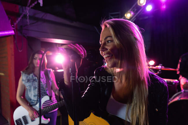 Vue de côté gros plan d'une chanteuse caucasienne aux longs cheveux blonds jouant dans un lieu musical avec une guitare acoustique, tenant le microphone et souriant, une bassiste sur scène en arrière-plan et des projecteurs roses — Photo de stock