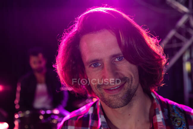 Ritratto di un musicista caucasico sul palco durante una permanenza in un locale musicale, sorridente alla telecamera sotto le luci rosa, con un batterista seduto su un kit di batteria sullo sfondo — Foto stock