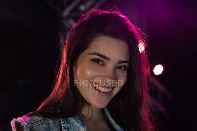Retrato de uma atraente músico feminino caucasiano com longos cabelos escuros no palco durante uma permanência em um local de música, sorrindo para a câmera sob luzes rosa — Fotografia de Stock