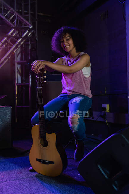 Retrato de una mujer de raza mixta sentada en un taburete de tambor, sosteniendo una guitarra acústica, mirando a la cámara y sonriendo mientras instala equipo de banda en el escenario en un lugar de música antes de una actuación - foto de stock
