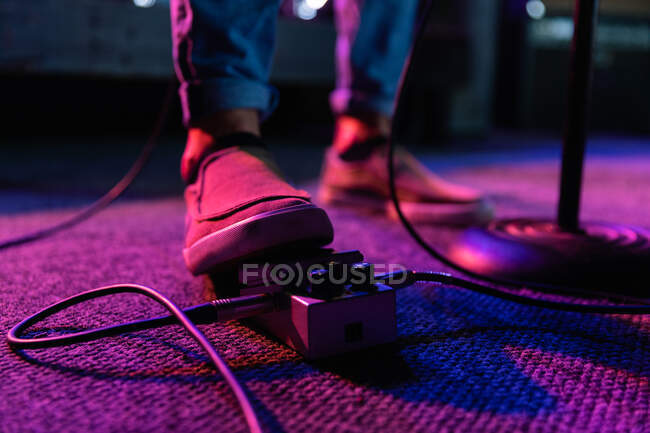 Vista frontal de sección baja detalle del pie de un guitarrista tocando en un escenario iluminado utilizando un pedal de efectos para cambiar su sonido de guitarra mientras actúa con una banda en un lugar de música, bajo luces rosadas - foto de stock