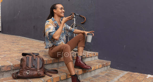Vista lateral de um homem de raça mista com longos dreadlocks para fora e sobre a cidade em um dia ensolarado, sentado na rua e sorrindo, usando um smartphone e segurando uma xícara de café, com sua bicicleta encostada à parede ao lado dele . — Fotografia de Stock