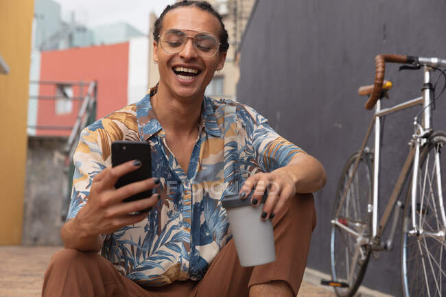 Vista frontal de cerca de un hombre de raza mixta con largas rastas en la ciudad en un día soleado, sentado en la calle y sonriendo, usando un teléfono inteligente y sosteniendo una taza de café, con su bicicleta de pie junto a él. - foto de stock