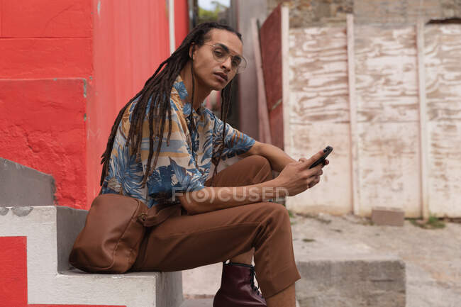 Вид сбоку на человека смешанной расы с длинными ногами в солнечный день, сидящего на лестнице на улице и использующего смартфон, глядя прямо в камеру. — стоковое фото