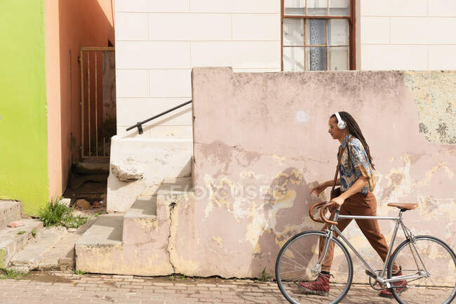 Vue latérale d'un homme de race mixte avec de longs dreadlocks dehors et autour dans la ville par une journée ensoleillée, portant des écouteurs, marchant dans la rue et faisant rouler son vélo. — Photo de stock