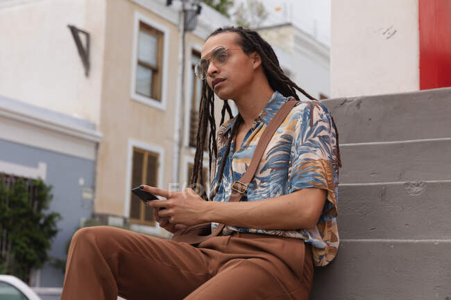 Front view close up of a mixed race man with long dreadlocks out and about in the city on a sunny day, sitting on the stairs in the street and using a smartphone, looking straight into camera. — Stock Photo