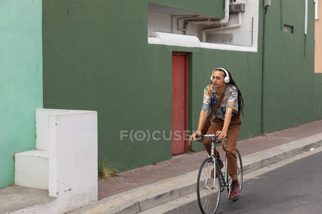 Вид спереди на смешанного расового человека с длинными дредами в городе в солнечный день, в наушниках, катающегося на велосипеде по улице. — стоковое фото