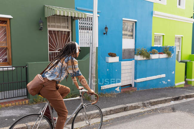 Visão traseira de um homem de raça mista com longos dreadlocks para fora e sobre a cidade em um dia ensolarado, usando fones de ouvido, andando de bicicleta na rua . — Fotografia de Stock
