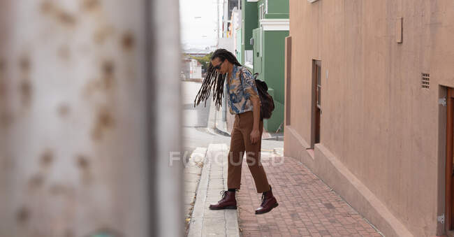 Vista lateral de um homem de raça mista com longos dreadlocks para fora e sobre a cidade em um dia ensolarado, vestindo uma mochila e óculos de sol, andando na rua . — Fotografia de Stock