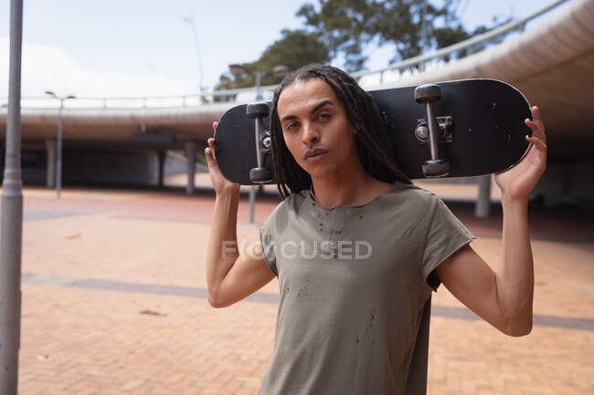 Front view of a mixed race man with long dreadlocks out and about in the city on a sunny day, standing in the street next to a bridge, holding a skateboard and looking straight into camera. — Stock Photo