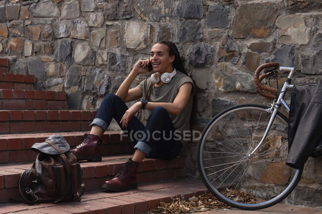 Vista lateral de un hombre de raza mixta con largas rastas en la ciudad en un día soleado, sentado en las escaleras de la calle y sonriendo, usando un teléfono inteligente, con su bicicleta apoyada contra la pared a su lado. - foto de stock