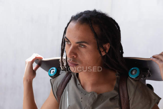 Front view close up of a mixed race man with long dreadlocks out and about in the city on a sunny day, holding a skateboard. — Stock Photo