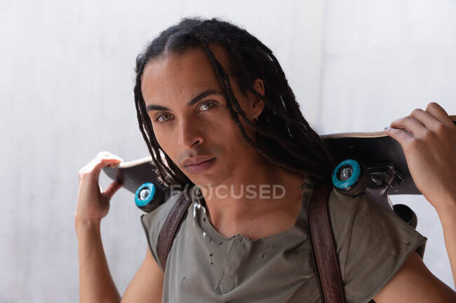 Front view close up of a mixed race man with long dreadlocks out and about in the city on a sunny day, holding a skateboard and looking straight into camera. — Stock Photo