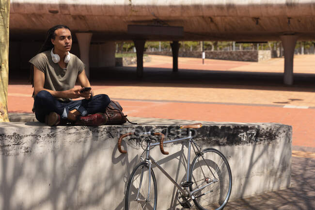 Вид сбоку на смешанного гонщика с длинными дредами в городе в солнечный день, сидящего на стене на улице и, используя смартфон, с велосипедом, прислоненным к стене рядом с ним. — стоковое фото