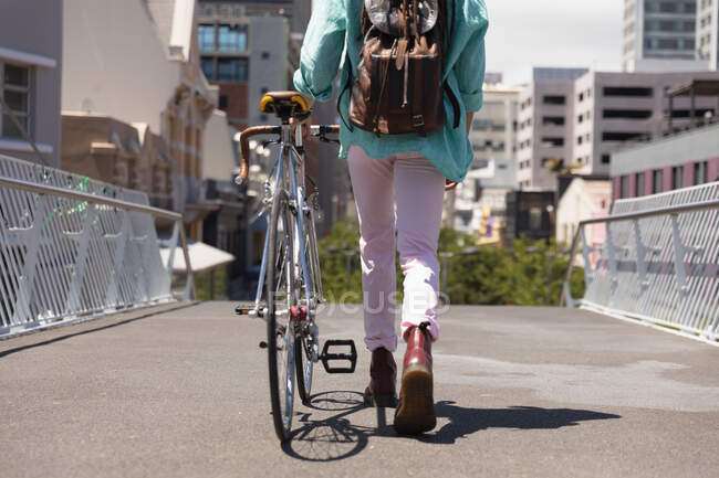 Visão traseira seção baixa do homem com longos dreadlocks para fora e sobre a cidade em um dia ensolarado, usando mochila, andando na rua e rodando sua bicicleta . — Fotografia de Stock