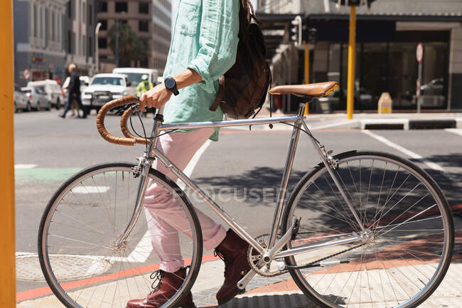 Vista laterale bassa sezione dell'uomo con lunghi dreadlocks in giro per la città in una giornata di sole, indossando lo zaino, camminando per strada e ruote la sua bicicletta. — Foto stock