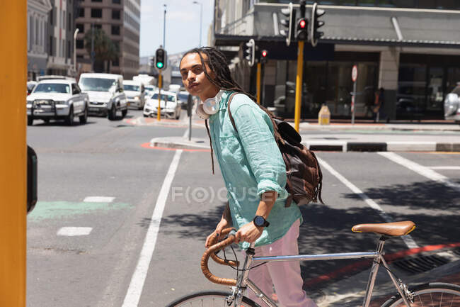 Vue latérale d'un homme de race mixte avec de longs dreadlocks dehors et dans la ville par une journée ensoleillée, portant un sac à dos, marchant dans la rue et roulant son vélo. — Photo de stock